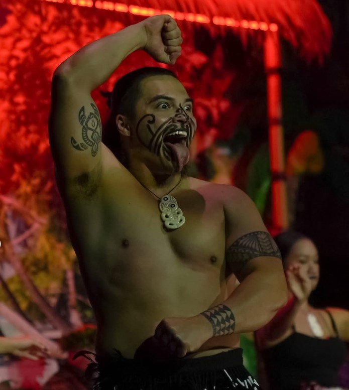 Person performing traditional dance, with face paint and tattoos, sticking out tongue.