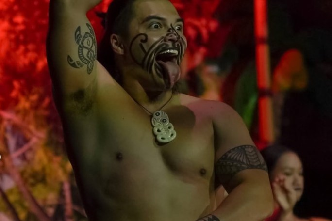 Person performing traditional dance, with face paint and tattoos, sticking out tongue.