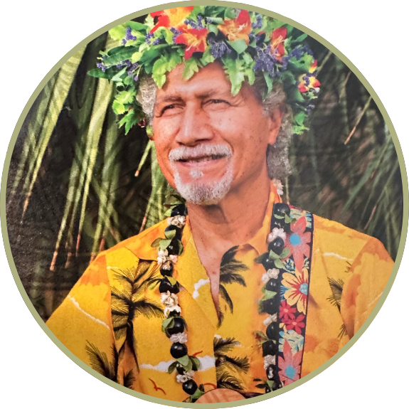 Smiling man in colorful floral attire and head lei against a leafy background.