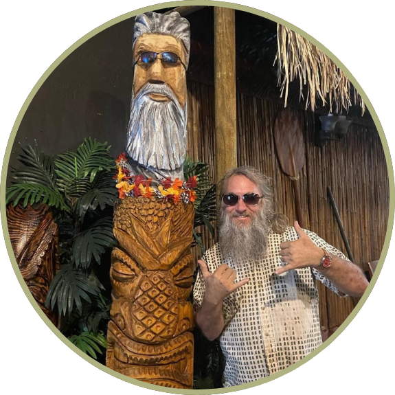 Man with sunglasses poses next to a tiki statue with a floral lei and tropical decor.
