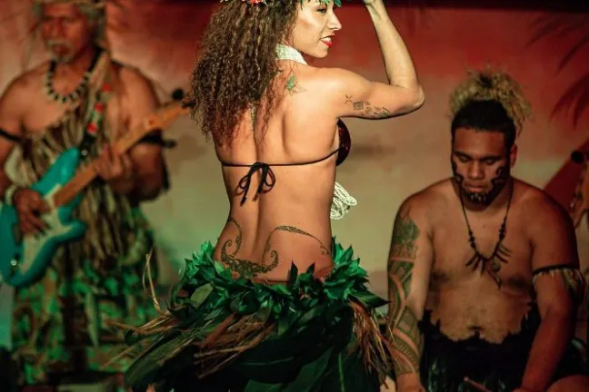 Hula dancer in floral skirt and headpiece performing with musicians in traditional attire playing instruments.