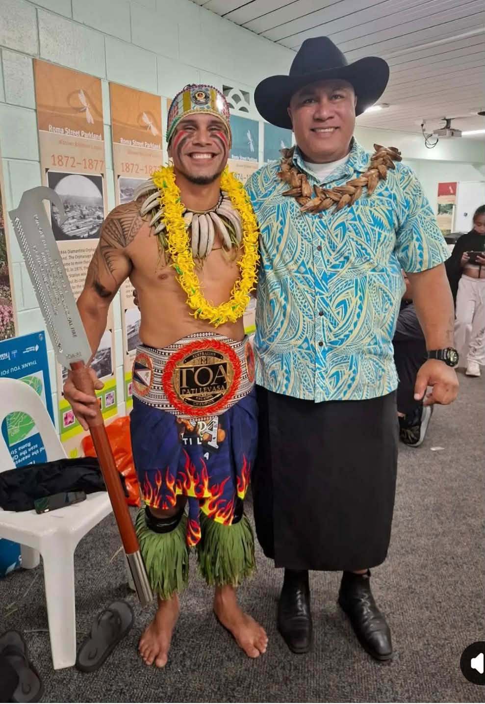 Two men in Samoan attire, one with a lei and staff, both smiling indoors.